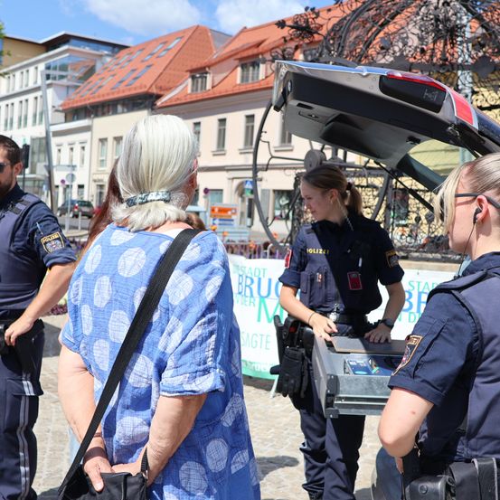 Eine Polizistin benutzt ein Gerät im Kofferraum eines Autos, während eine weitere Polizistin in der Nähe steht und zwei Frauen beobachten.