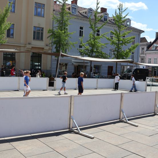 Mehrere Kinder spielen auf einem provisorischen Spielfeld, das von Barrieren umgeben ist, in einer urbanen Umgebung mit Gebäuden und Bäumen im Hintergrund.