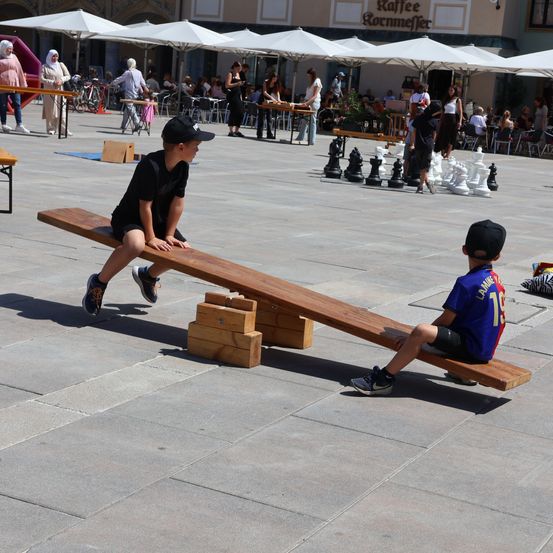 Zwei Jungen spielen auf einem Wippbrett in einer Freiluft-Plaza, umgeben von Schachbrettern und Menschen, die an Tischen sitzen.