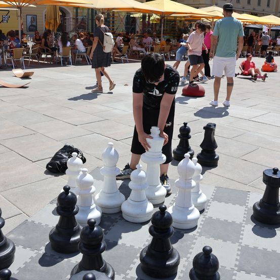 Ein Mann spielt Schach auf einem großen Brett im Freien auf einem öffentlichen Platz, wo Leute herumlaufen.