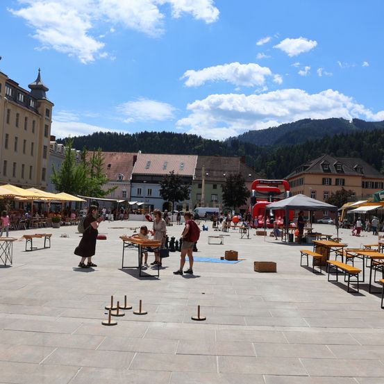 Ein geschäftiger Stadtplatz mit Menschen, die Schach spielen, umgeben von Gebäuden und Bergen im Hintergrund.