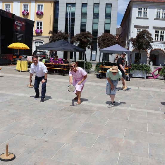 Drei Personen spielen ein Kegelspiel auf einem gepflasterten Platz, mit einem gelben Tisch und Zelten im Hintergrund.