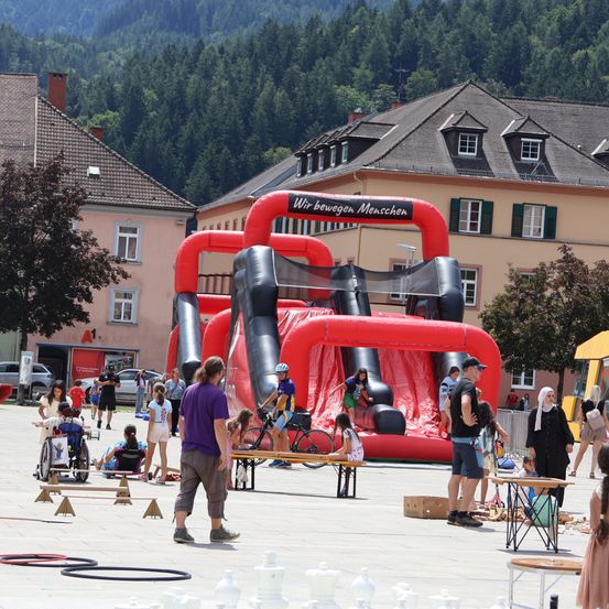 Eine Gruppe von Menschen versammelt sich um einen aufblasbaren Hindernisparcours auf einem Stadtplatz, mit Bergen im Hintergrund. Einige spielen auf dem Parcours, während andere zuschauen.