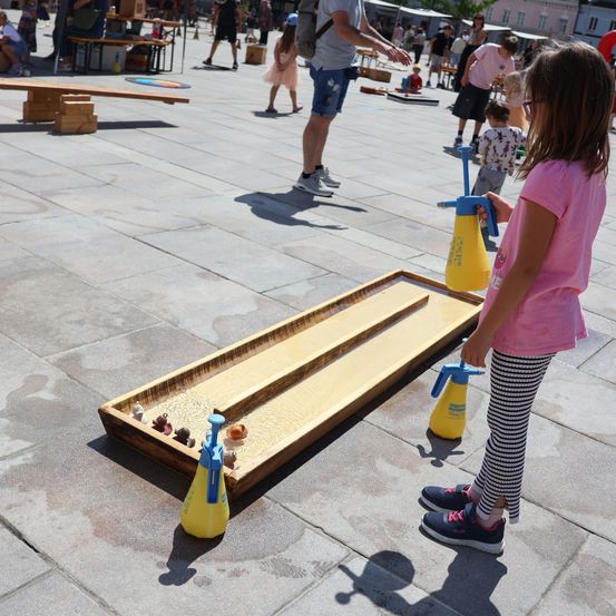 Ein junges Mädchen spielt ein wasserbasiertes Spiel auf einem Holzbrett auf einem öffentlichen Platz, mit anderen Kindern und Erwachsenen in der Umgebung.
