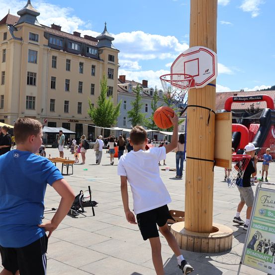 Ein Junge in einem weißen T-Shirt und schwarzen Shorts spielt Basketball an einem Holzpfahl auf einem Stadtplatz mit anderen Leuten um ihn herum.