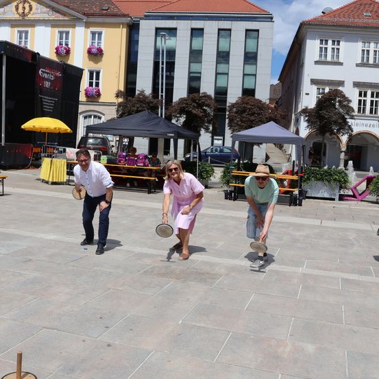 Drei Personen spielen ein Cornhole-Spiel auf einem gepflasterten Außenbereich. Die Frau in der Mitte trägt ein rosa Kleid. Hinter ihnen befinden sich Gebäude, Zelte und Bäume.