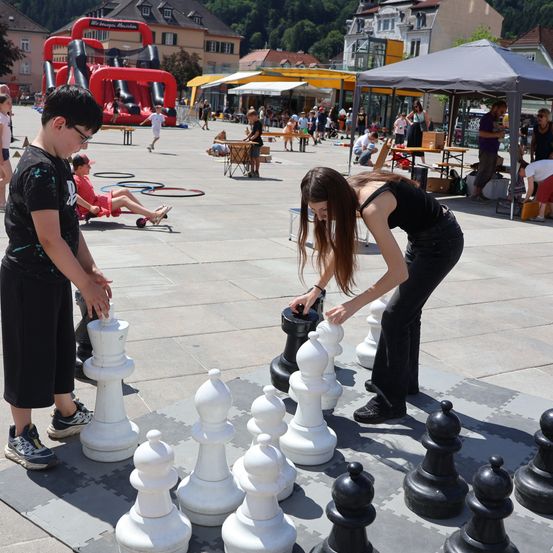 Zwei Kinder spielen Schach auf einem großen Brett in einer öffentlichen Plaza, mit einem Hüpfburg und anderen Aktivitäten im Hintergrund.