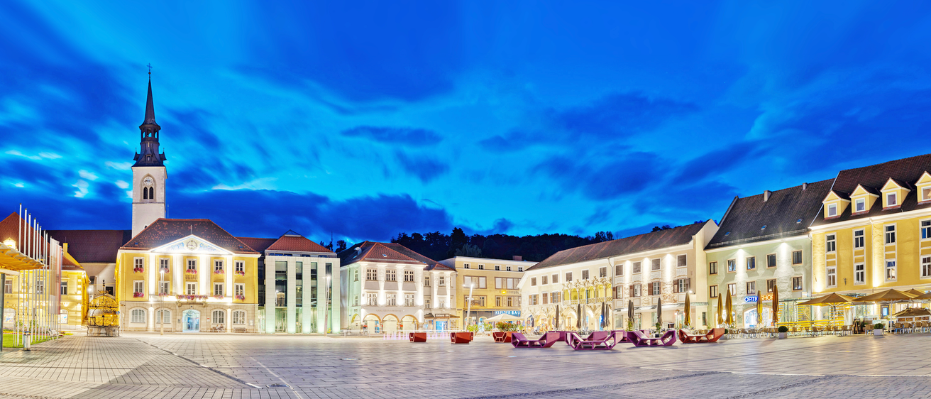 Eine Stadtlandschaft mit modern beleuchteten Gebäuden bei Nacht unter einem blauen Himmel mit Wolken, umgeben von lila Stühlen und dekorativen Lichtern.