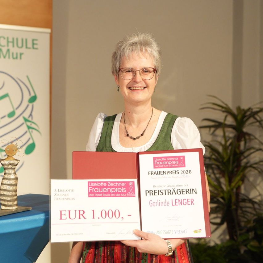 Eine Frau in traditioneller Kleidung hält ein Zertifikat und einen Geldpreis und lächelt. Ein Pokal und eine Pflanze sind im Hintergrund.