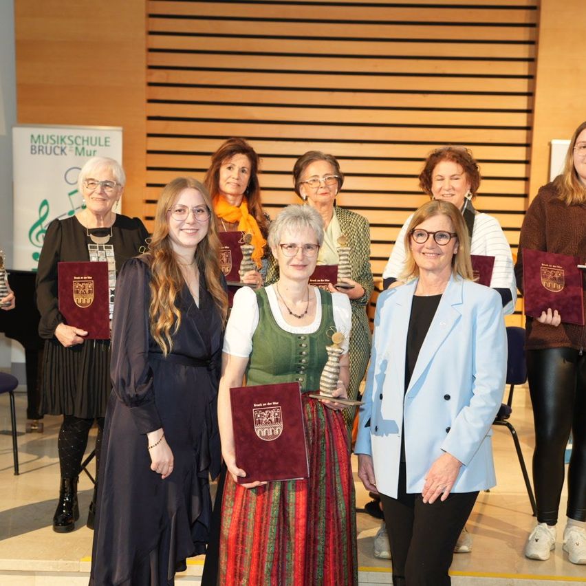 Eine Gruppe von Frauen steht zusammen auf einer Bühne und hält jeweils eine rote Auszeichnung. Sie lächeln und posieren für ein Foto. Die Frau in der Mitte trägt eine Brille und eine Halskette. Hinter ihnen steht eine Holzwand mit einem Schild, auf dem 'Musikschule Bruck Mur' steht.