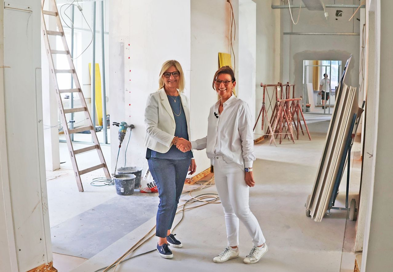 Zwei Frauen in formeller Kleidung stehen in einer Baustelle, lächeln und schütteln Hände. Hinter ihnen sind eine Leiter, Eimer und Baugeräte zu sehen. Eine weitere Person ist im Hintergrund zu sehen.
