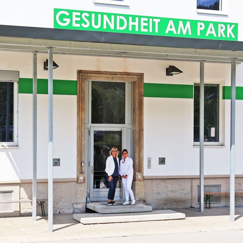 Zwei Frauen in weißen Outfits stehen vor einem Gebäude mit einem grünen Schild, das 'Gesundheit am Park' steht. Das Gebäude hat Glasfenster und Türen und ist von Metallpfosten umgeben.