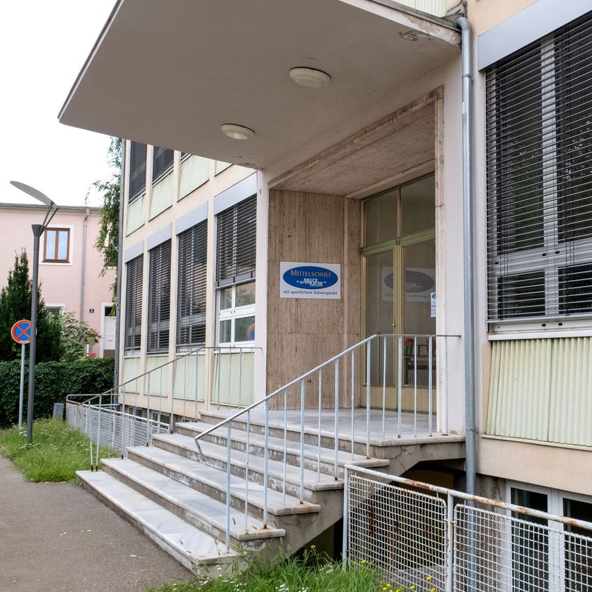 Der Eingang eines Gebäudes mit einem blauen Schild, das 'Mittelschule' steht, mit einer Treppe mit Handläufen, die zu einer Glastür führt, umgeben von einem kleinen Garten und einer Straßenlaterne.