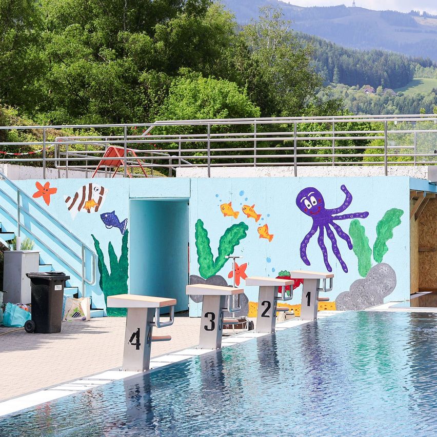 Ein Schwimmbecken mit Sprungbrettern, nummeriert von 1 bis 4, umgeben von einer blauen Wand mit Meerestieren und Bergen im Hintergrund.