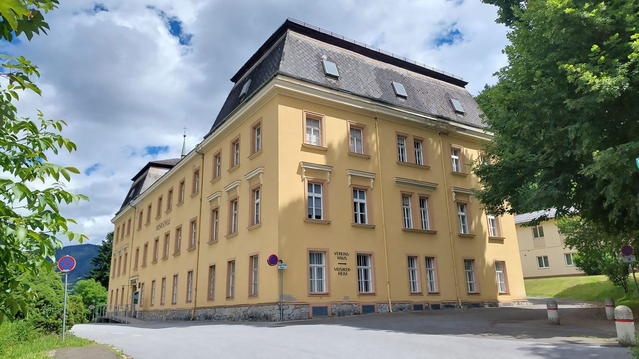 Ein großes gelbes Gebäude mit vielen Fenstern und einem grauen Dach, beschriftet mit 'Vereinshaus Musikverein Heim', unter einem bewölkten Himmel.
