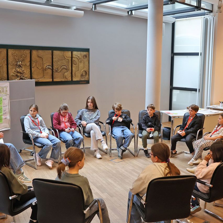 Eine Gruppe von Kindern sitzt in einem Klassenzimmer auf Stühlen, einige schauen zu einer Lehrerin, die vor ihnen steht. Das Klassenzimmer hat einen Holzboden und eine Tafel an der Wand.