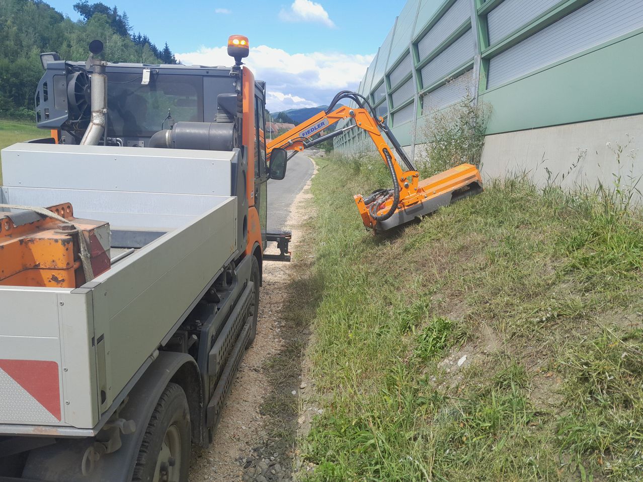 Ein großer orangefarbener Maschinenwagen ist am Straßenrand geparkt, wobei sein Arm ausgestreckt ist, um Gras in der Nähe eines Gebäudes zu schneiden.