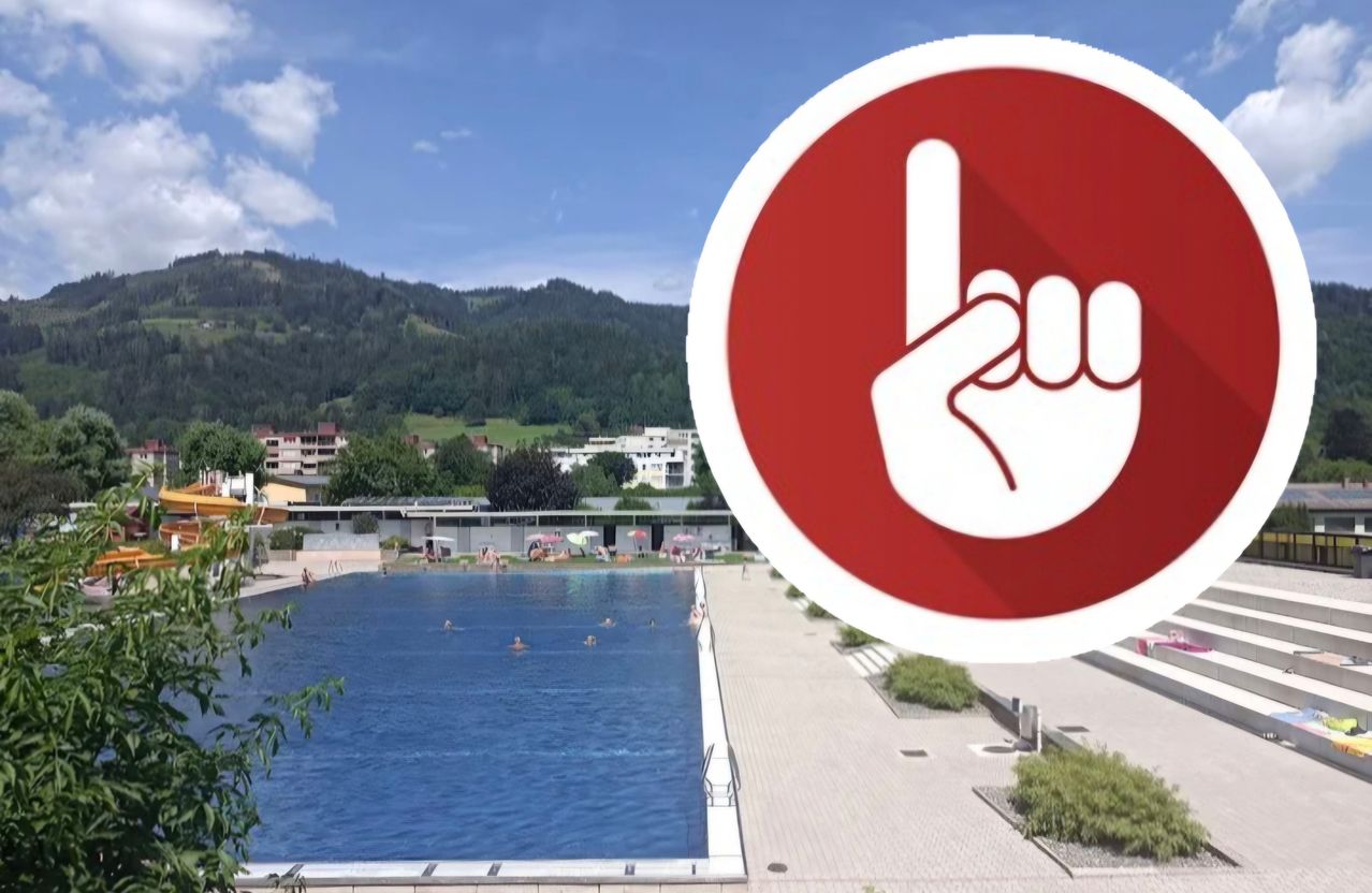 Freibad mit Schwimmenden, umgeben von Gebäuden und Grünflächen, mit einem Berg im Hintergrund.