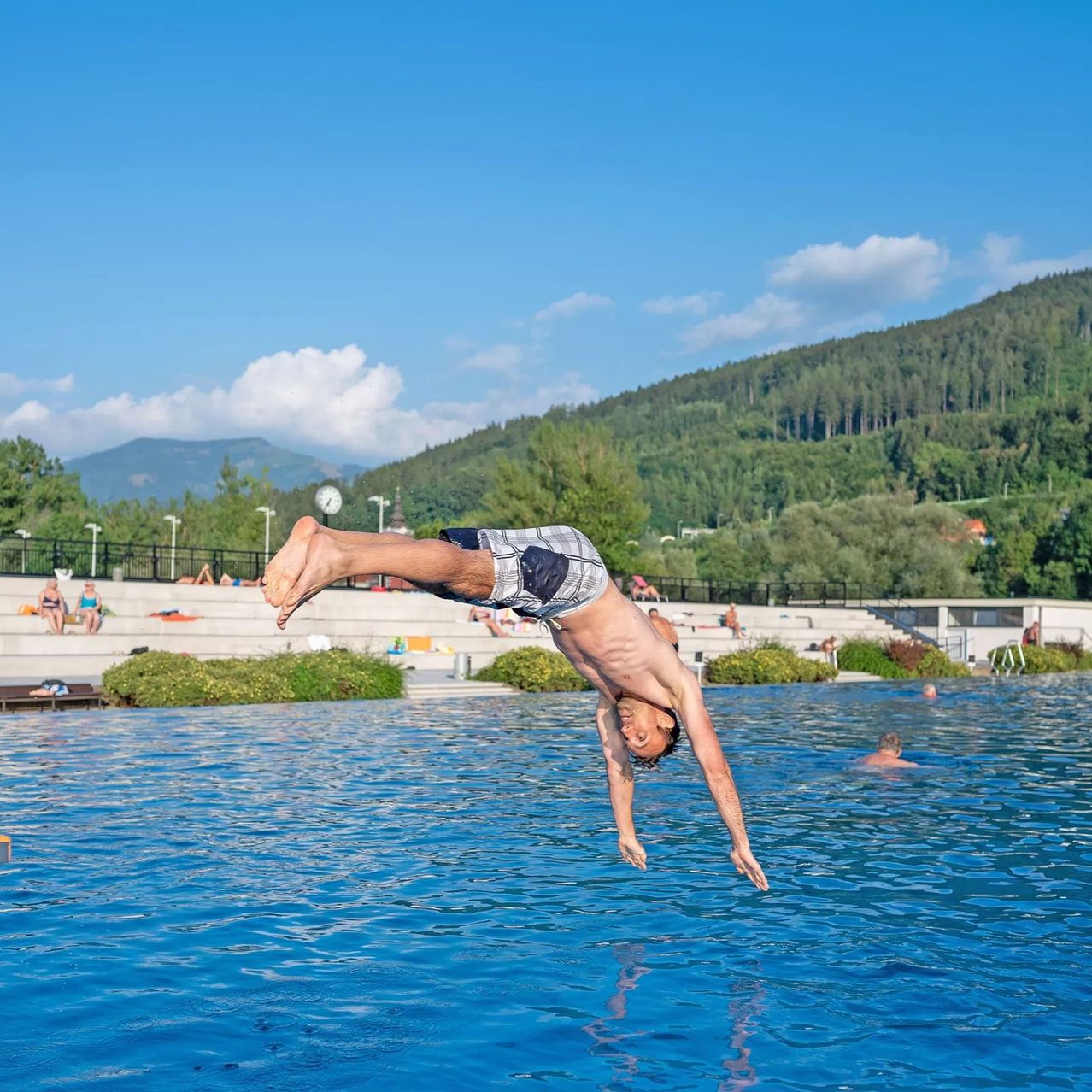 Ein junger Mann führt einen Handstand-Sprung in ein Schwimmbecken mit einer malerischen Bergkulisse aus.