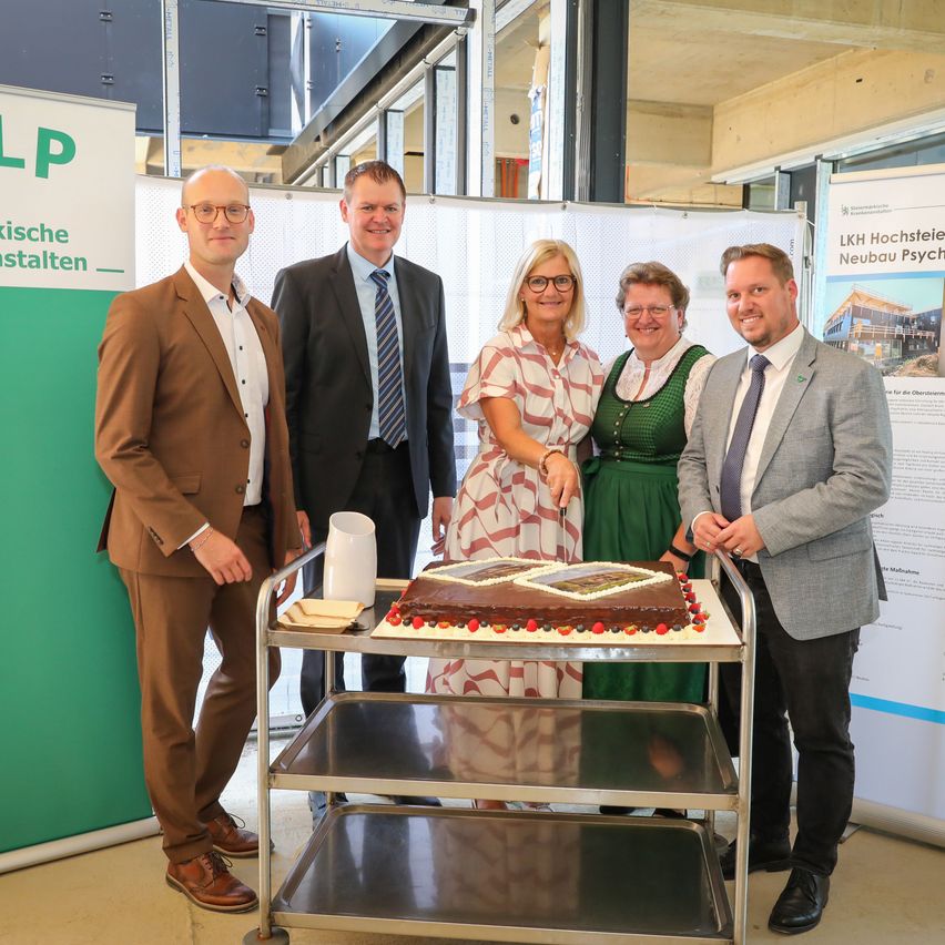 Fünf Personen stehen um einen Kuchen auf einem Wagen, zwei Frauen in der Mitte schneiden ihn an. Dahinter ist ein Banner mit dem Text 'LP' zu sehen.
