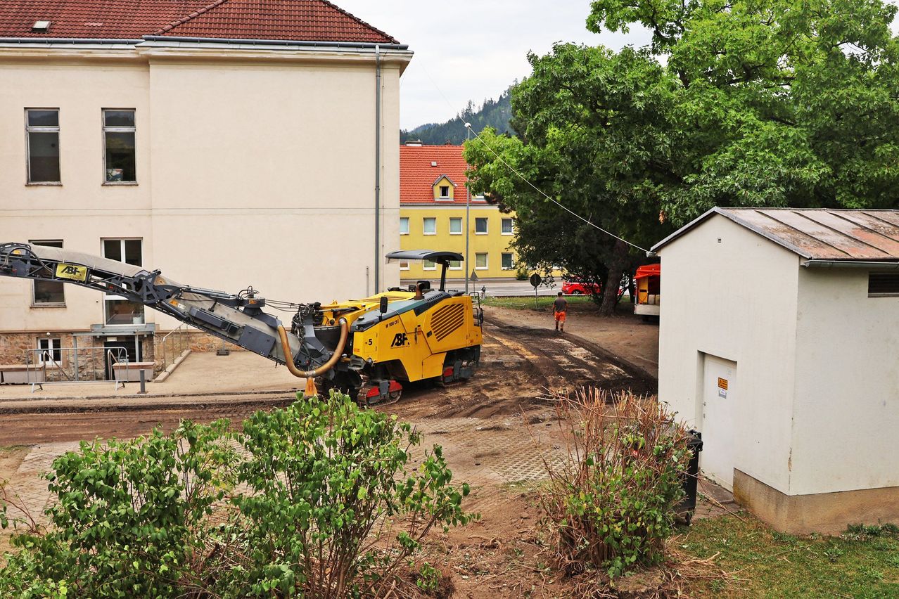 Ein gelber Baufahrzeug steht auf einer unbefestigten Straße in der Nähe eines Gebäudes mit rotem Dach. Eine Person in einem orangefarbenen Shirt geht auf der Straße.