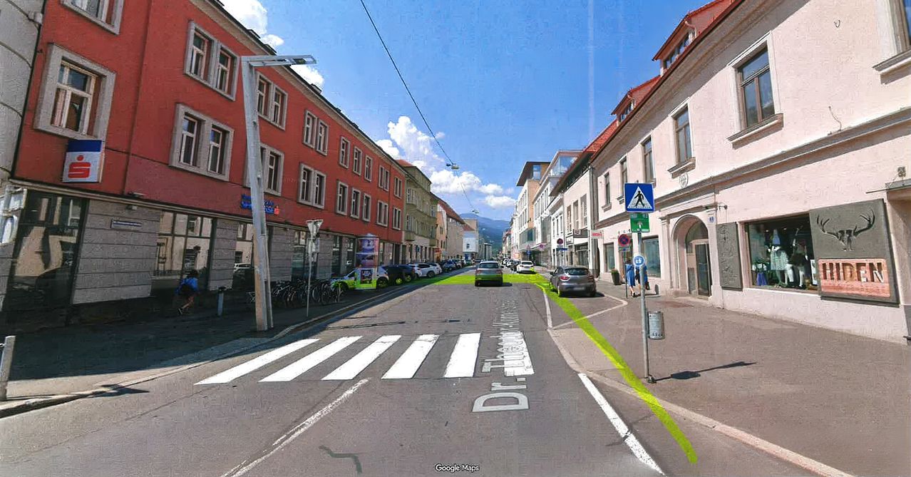 Eine Straßenansicht mit Gebäuden auf beiden Seiten, einem grünen Fahrradweg und einem Zebrastreifen. Autos sind am Straßenrand geparkt.