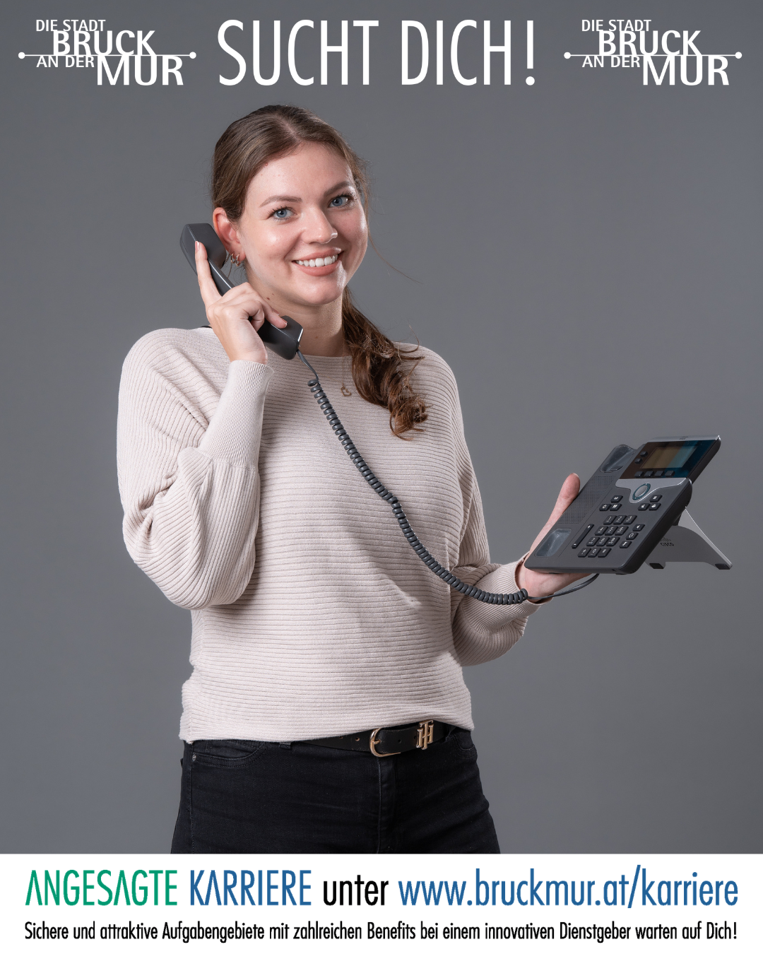 Eine Frau in einem beigen Pullover und schwarzen Hosen hält ein Telefon und eine Telefonbasis. Sie lächelt und schaut in die Kamera.