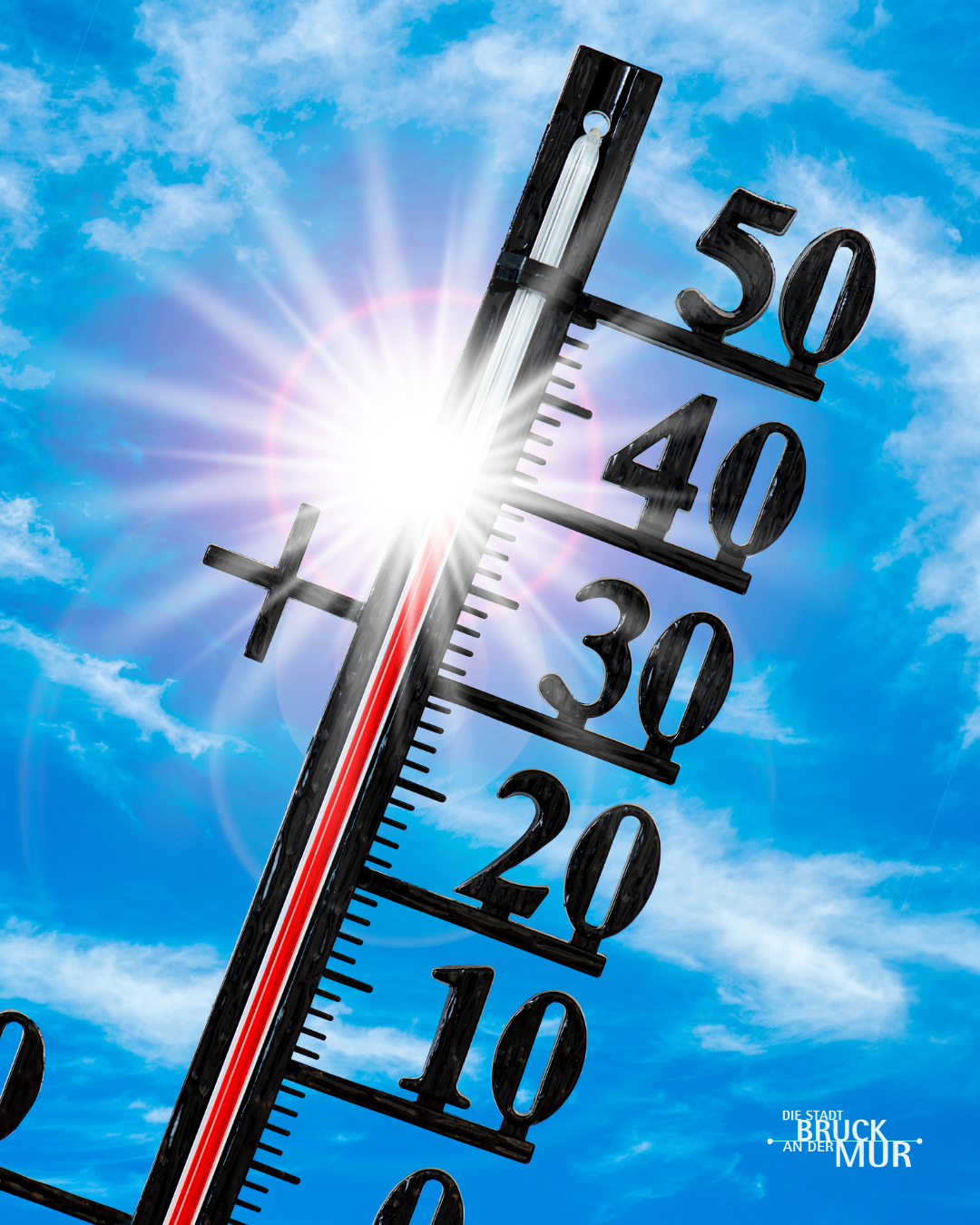 Ein Thermometer mit einer hohen Temperaturanzeige, Zahlen von 10 bis 50, unter einem hellen Sonnenschein gegen einen blauen Himmel mit Wolken.