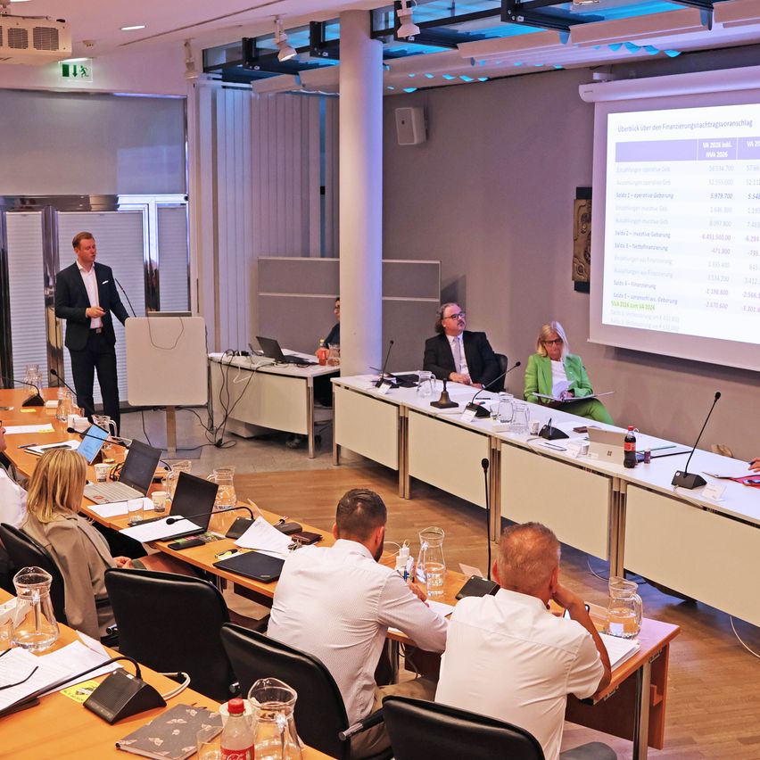 Ein Konferenzraum mit einem Mann, der am Rednerpult eine Präsentation hält. Das Publikum sitzt vor ihm. Eine Projektionsleinwand zeigt Finanzdaten an.