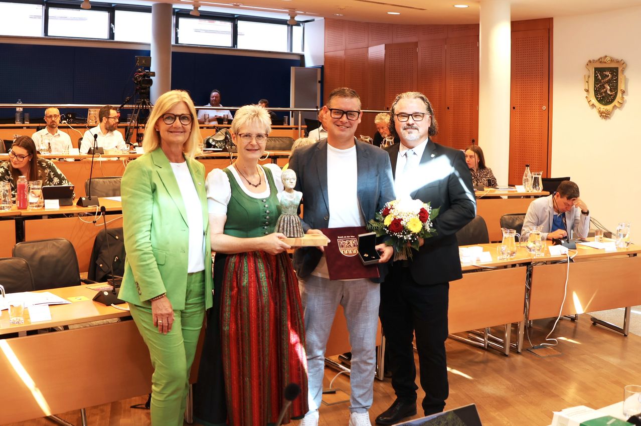 Drei Personen stehen in einer Innenumgebung, zwei Frauen und ein Mann, alle halten Auszeichnungen und Blumen. Hinter ihnen sitzen einige Personen auf Stühlen, und ein Mikrofon steht auf einem Ständer.