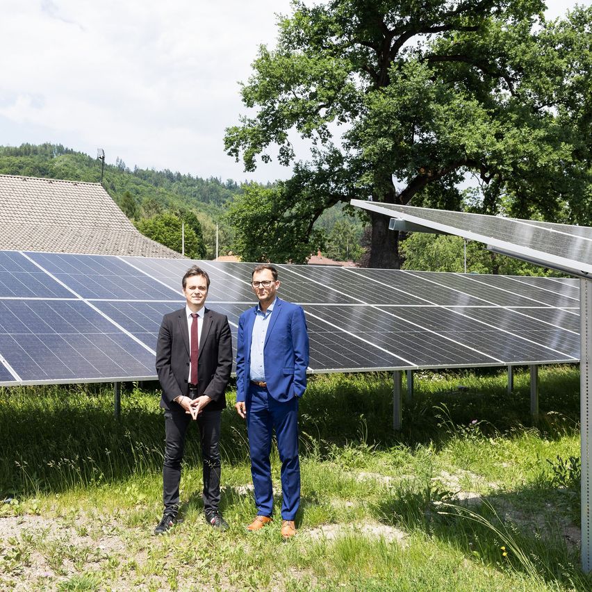 Zwei Männer stehen vor Solarpanelen in einem Grasfeld. Dahinter steht ein Haus und Bäume.