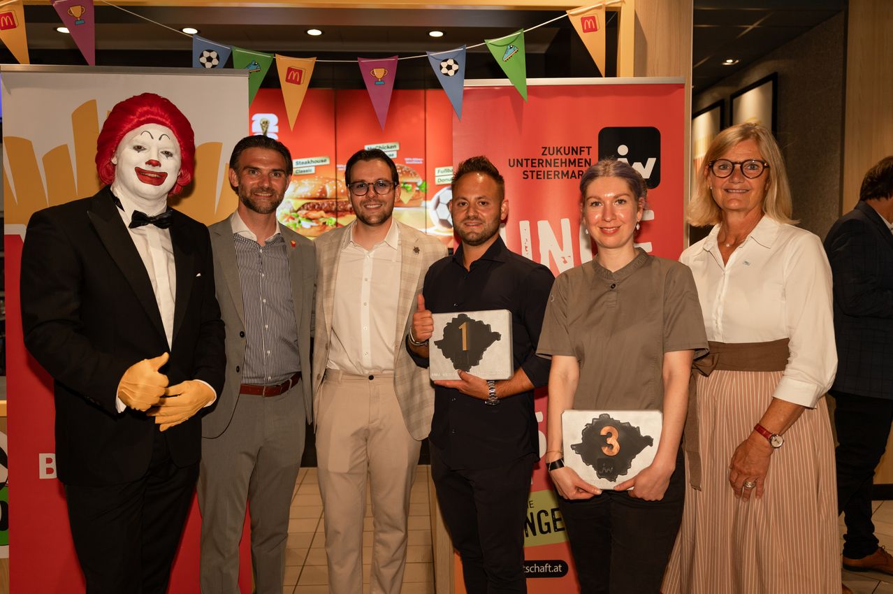 Five people stand next to each other, holding trophies. Two men wear glasses and suits. The woman in the middle holds a trophy with the number 3. Behind them is a banner with the text Zukunft Unternehmen Steiermark.