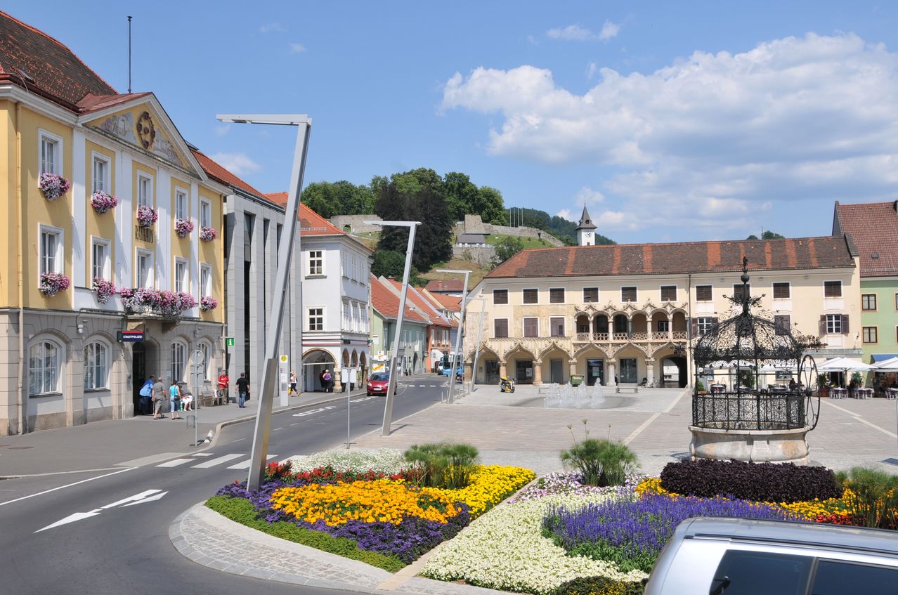 A town square with flower beds, street lamps, and buildings. People walk along the sidewalk, and a car is parked nearby. Trees and a castle are in the distance.