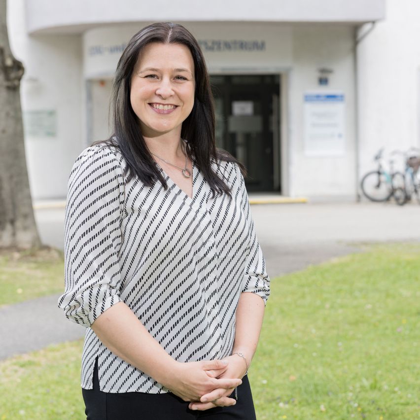 A smiling woman stands in front of a white building with the word Zentrum on it. She is wearing a white and black striped blouse. Behind her, there is a tree and two bicycles parked on the grass.