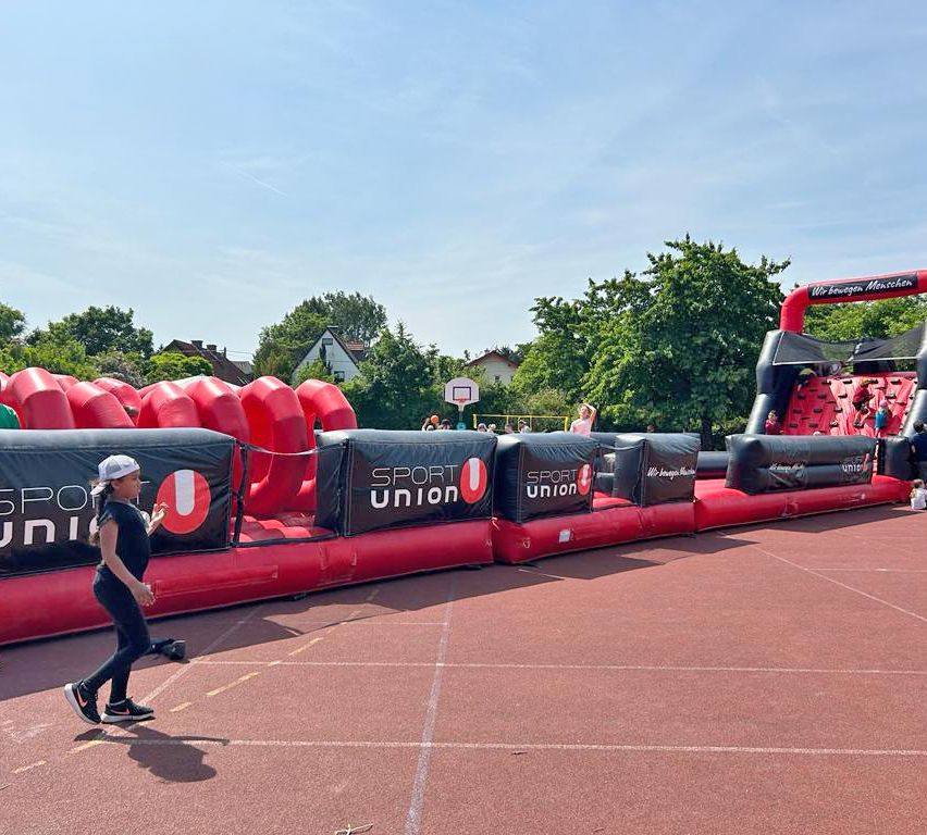 A woman runs on a track near a large inflatable obstacle course with red and black colors. Trees and a house are in the background.