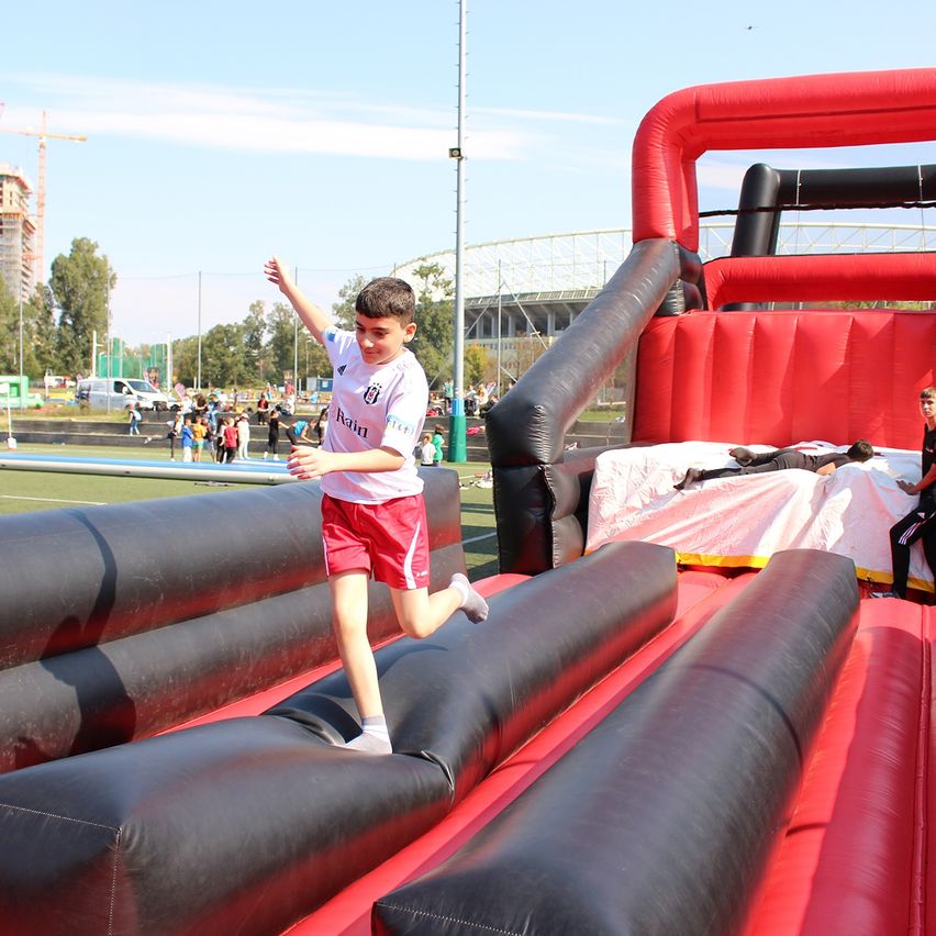 A boy wearing a white shirt runs on a bouncy castle at an outdoor event. Behind him, a person lies on a bed covered with a white cloth.