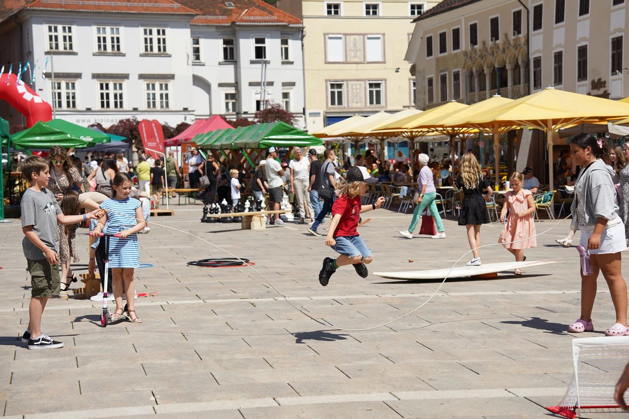 A young boy jumps over a rope while two girls hold it. Behind him, children play chess. Many people are around, some are sitting under umbrellas.