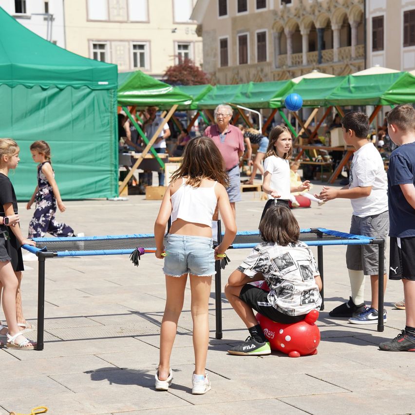 Kids play table tennis in an outdoor area. Several green tents are in the background. Some adults and children are walking or standing around.