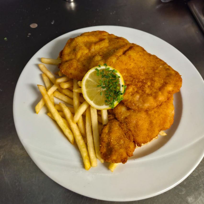 Ein weißer Teller mit gebratenem Schnitzel und Pommes, garniert mit einem Zitronenkeil und grünen Kräutern auf einer Metalloberfläche.