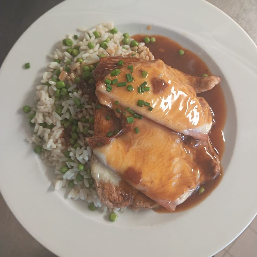 Ein Teller mit zwei Hähnchenbruststücken, die mit Sauce überzogen und mit grünen Zwiebeln garniert sind, serviert mit Reis und Erbsen.