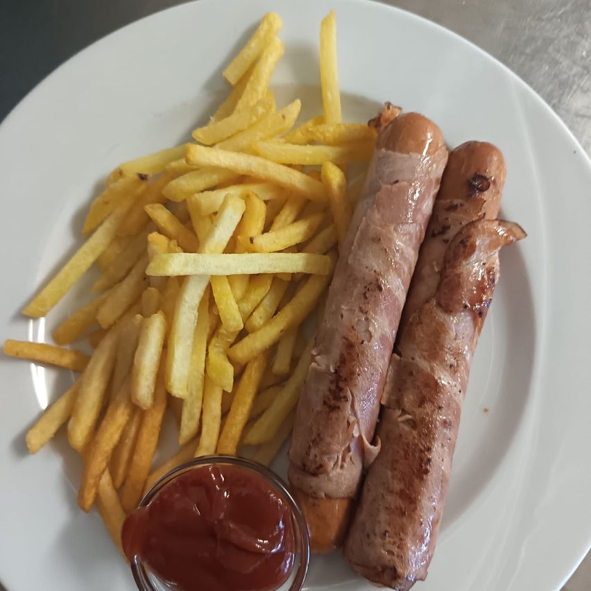 Ein weißer Teller mit zwei in Speck gewickelten Würstchen, golden-braunen Pommes und einem kleinen Behälter mit Ketchup.