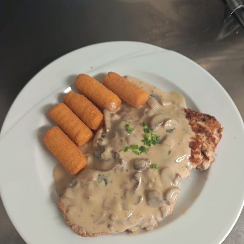 A white plate with a piece of chicken covered in creamy mushroom sauce and garnished with green herbs, accompanied by a side of golden cheese-filled sticks.