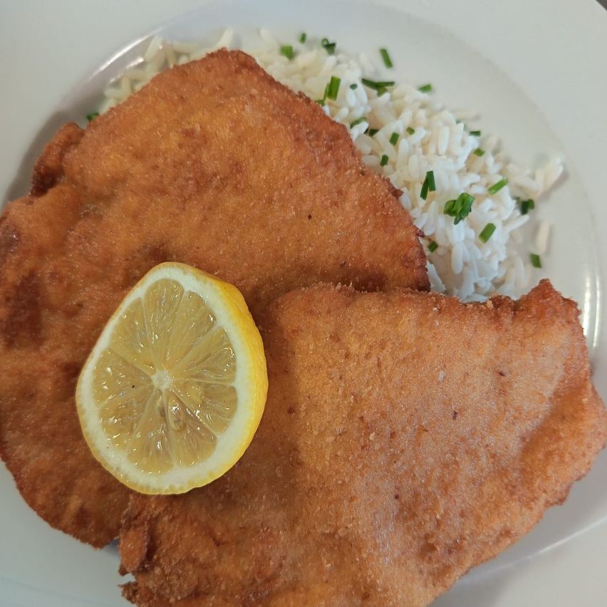 Gebratenes, paniertes Fleisch mit Zitronenscheibe, serviert mit weißem Reis und mit Schnittlauch bestreut auf einem weißen Teller.