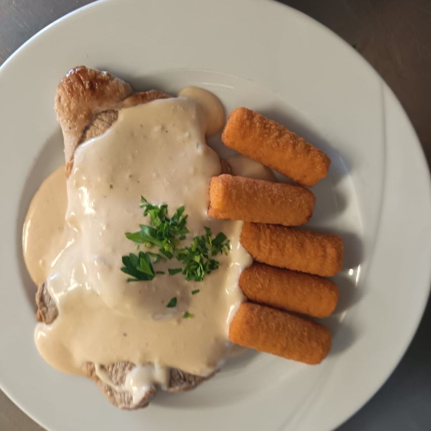 Ein weißer Teller mit einem Gericht, das ein Stück Fleisch mit cremiger Sauce, garniert mit grünen Kräutern, und begleitet von drei frittierten Stäbchen enthält.