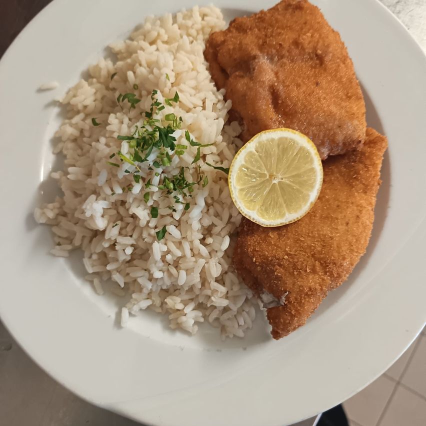Ein Teller mit Reis, gebratenem Fisch und einer Zitronenscheibe darauf. Der Reis ist mit grünen Kräutern garniert.