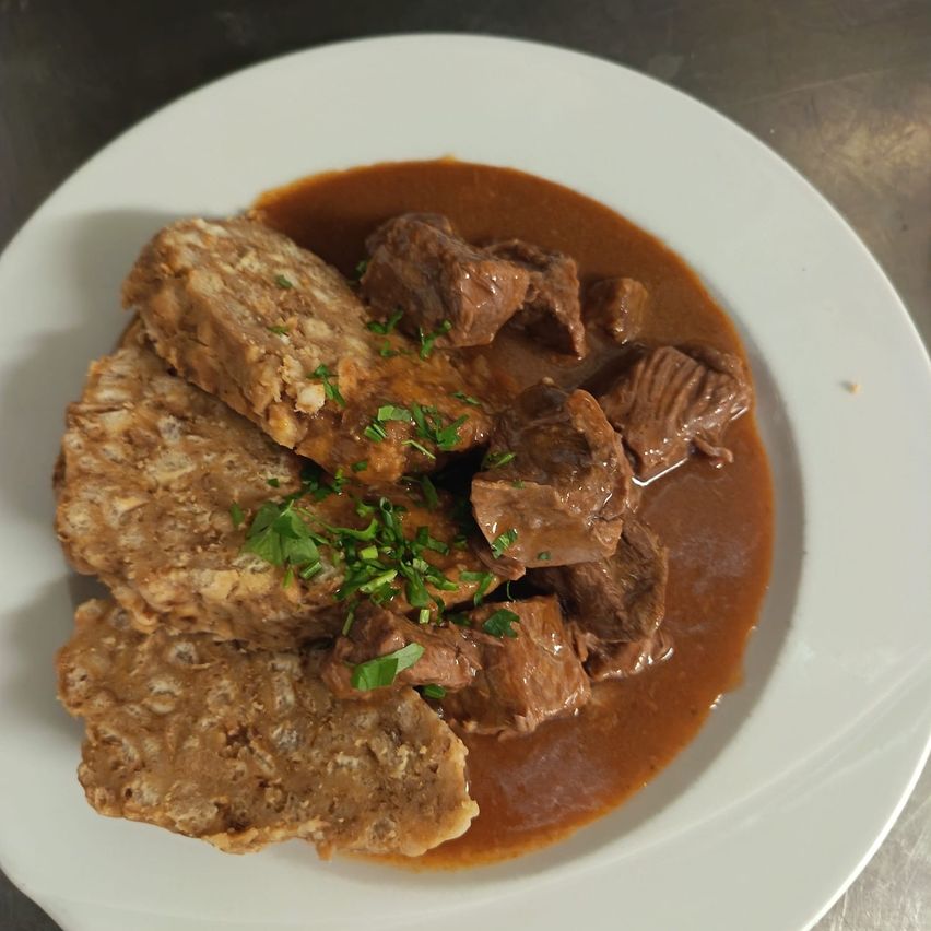 A plate of meat and gravy, topped with parsley, served on a white plate with a metal background.