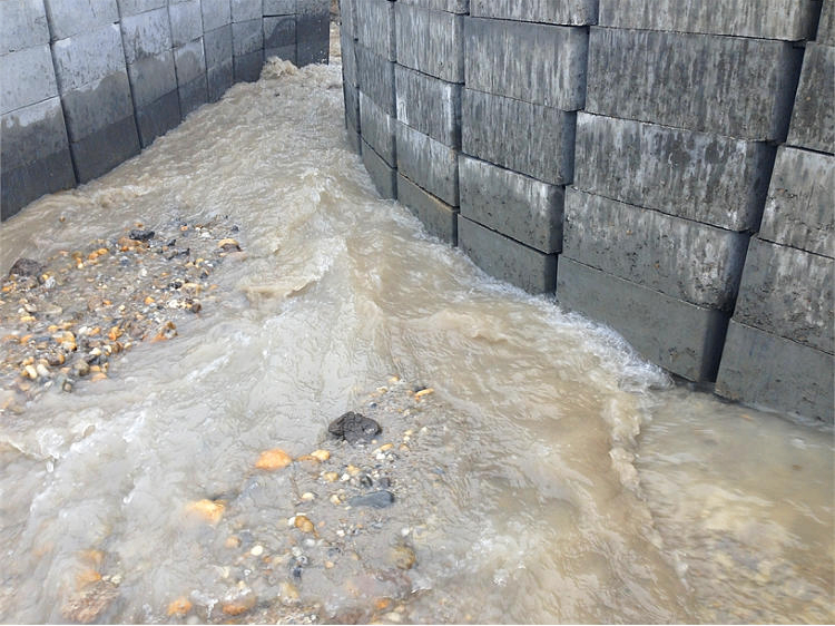 Eine Betonmauer leitet Wasser durch einen engen Spalt, was zu schnellem Fluss und Ansammlung von Trümmern führt.