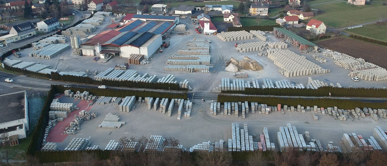 Luftaufnahme eines Industriegebiets mit einer großen Fabrik und Lagerhäusern, umgeben von Häusern und Feldern, mit Bergen im Hintergrund.