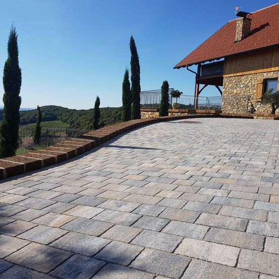 Eine Steinauffahrt führt zu einem Haus mit rotem Dach, umgeben von einem Zaun und hohen Bäumen, mit Blick auf Hügel und einen klaren blauen Himmel.