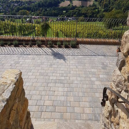 Eine gepflasterte Terrasse mit Metallgeländer bietet Blick auf Weinberge und die Landschaft. Schatten fallen auf den Weg.